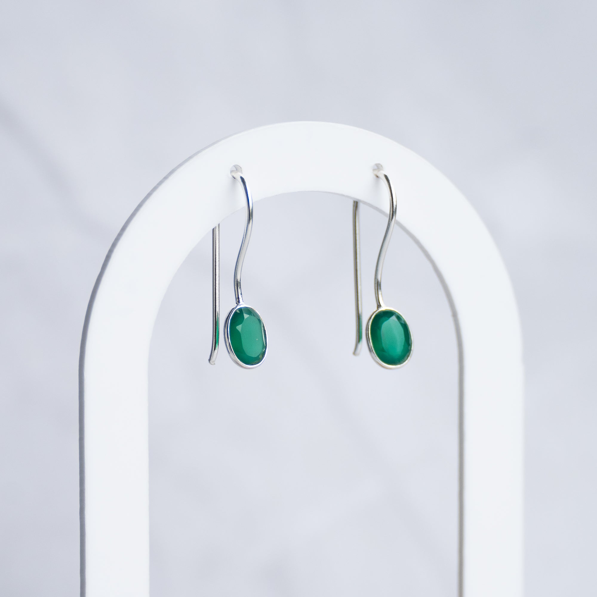A pair of silver drop earrings with oval green cabochon stones, hanging from a white arched jewelry display stand against a light gray marble background.