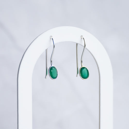 A pair of silver drop earrings with oval green cabochon stones, hanging from a white arched jewelry display stand against a light gray marble background.