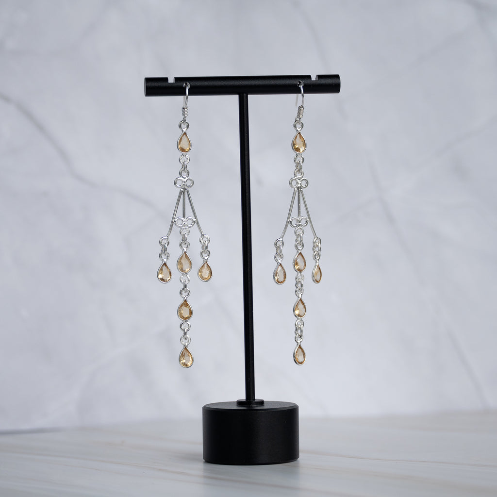 A pair of silver-toned dangle earrings with clear faceted beads and amber teardrop crystals, displayed on a black T-stand against a light gray marble background.