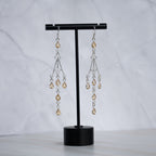 A pair of silver-toned dangle earrings with clear faceted beads and amber teardrop crystals, displayed on a black T-stand against a light gray marble background.