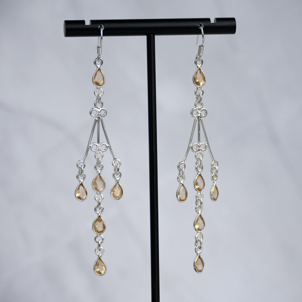 A pair of silver-toned dangle earrings with teardrop-shaped amber-colored crystals, displayed on a black T-stand against a light gray background.