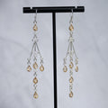 A pair of silver-toned dangle earrings with teardrop-shaped amber-colored crystals, displayed on a black T-stand against a light gray background.