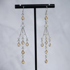 A pair of silver-toned dangle earrings with teardrop-shaped amber-colored crystals, displayed on a black T-stand against a light gray background.