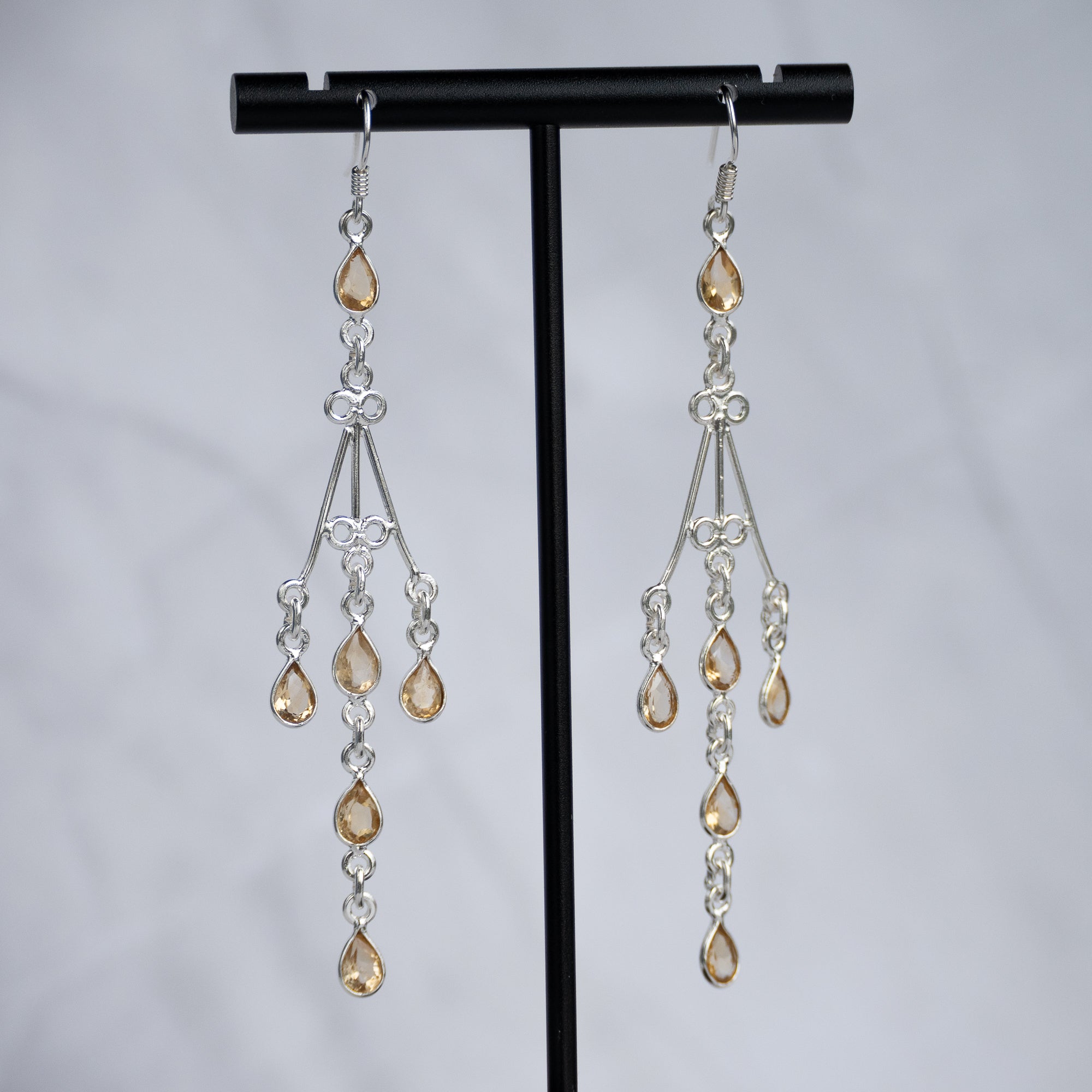 A pair of silver-toned dangle earrings with teardrop-shaped amber-colored crystals, displayed on a black T-stand against a light gray background.