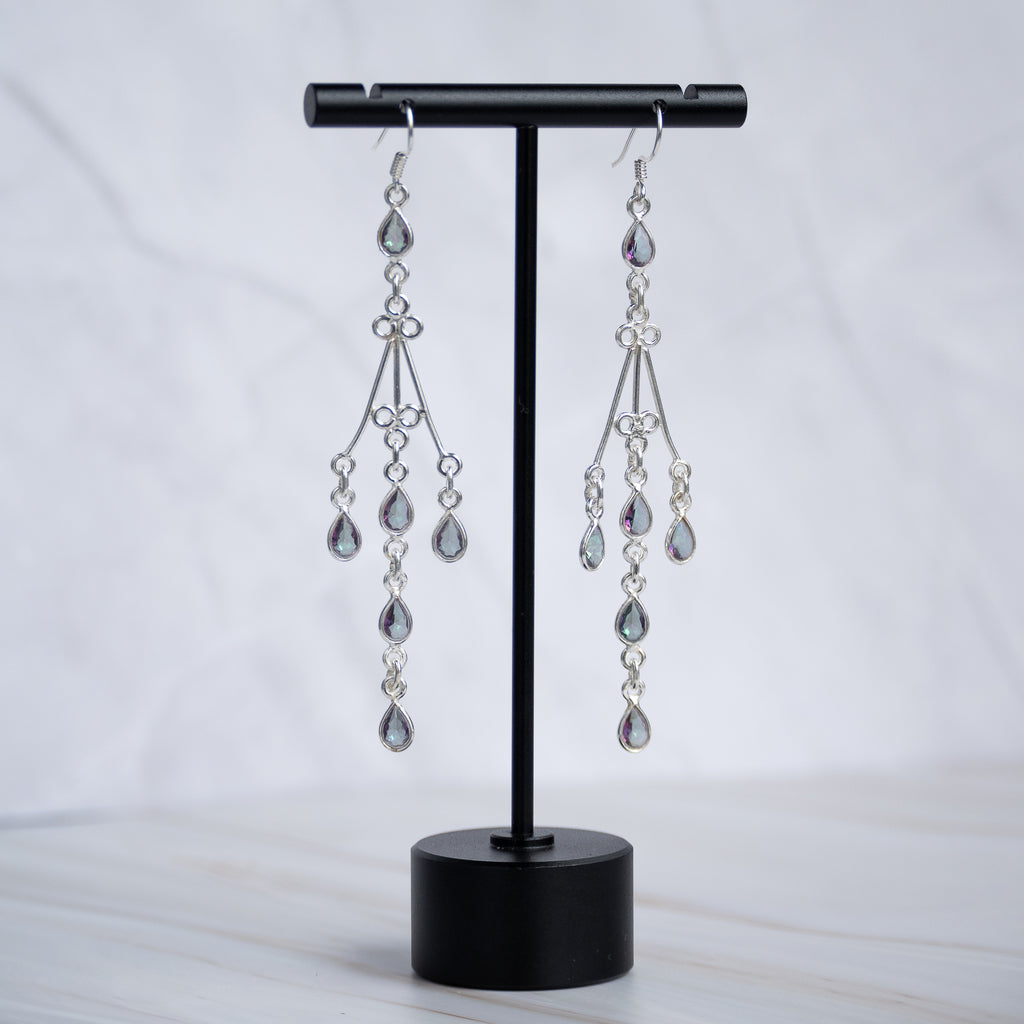 A pair of silver-toned dangle earrings with teardrop-shaped clear crystals, displayed on a black jewelry stand against a light marble background.