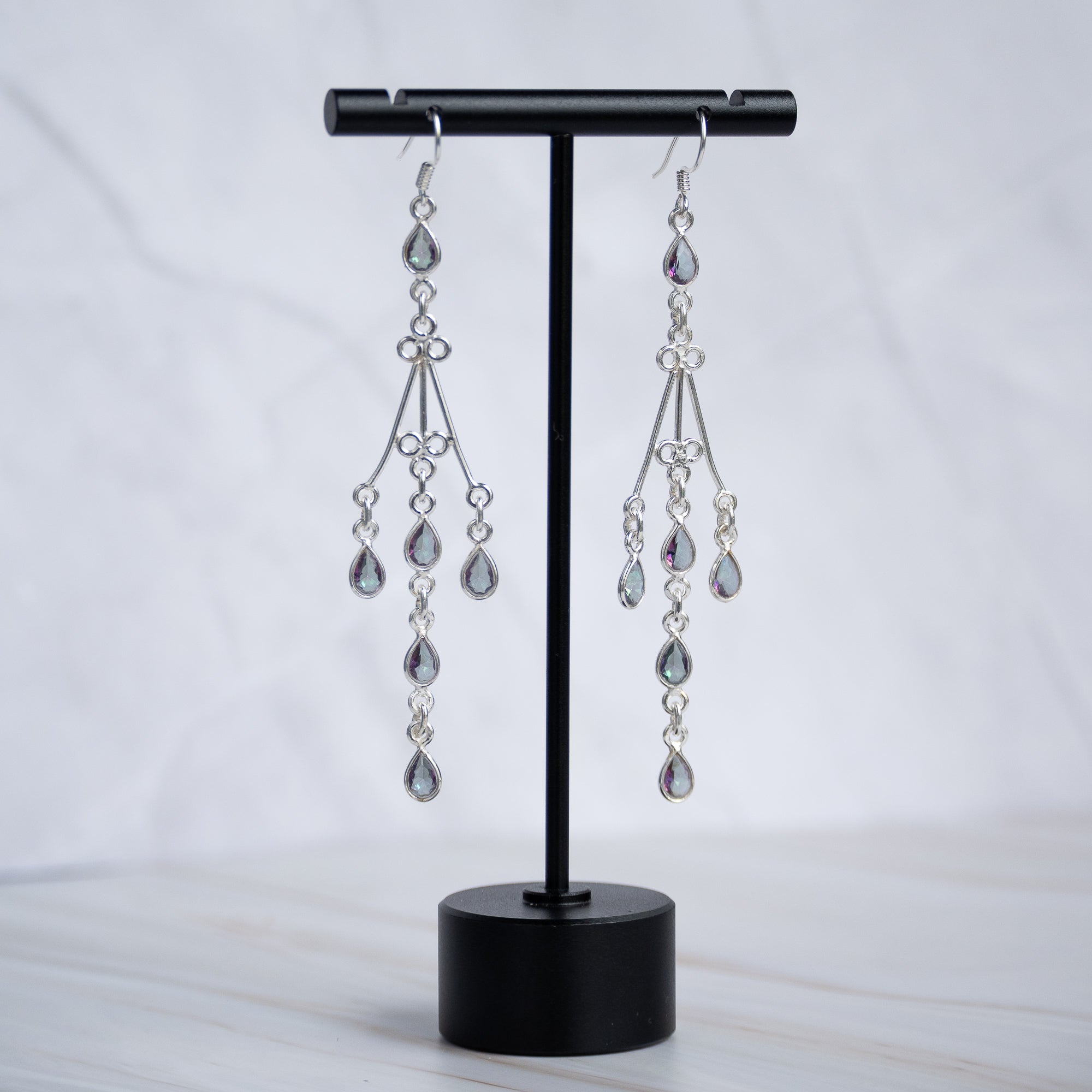 A pair of silver-toned dangle earrings with teardrop-shaped clear crystals, displayed on a black jewelry stand against a light marble background.
