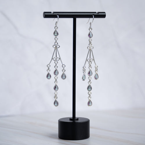 A pair of silver-toned dangle earrings with teardrop-shaped clear crystals, displayed on a black jewelry stand against a light marble background.