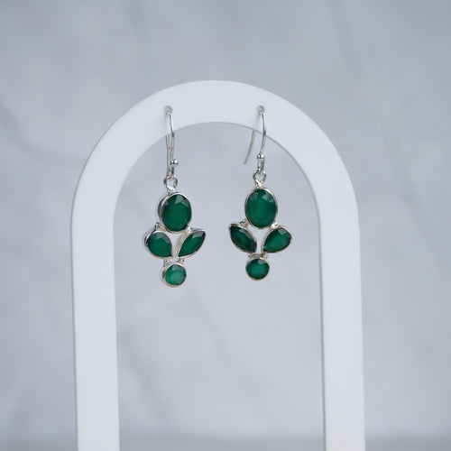 A pair of silver dangle earrings with green gemstone floral motifs, displayed on a white arched stand against a light gray background.