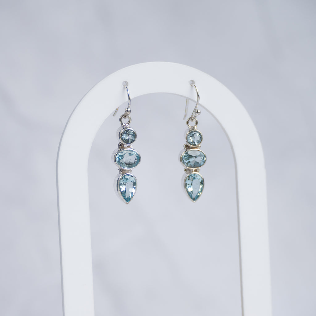 A pair of silver drop earrings with light blue gemstones, displayed on a white arched stand against a light gray background.