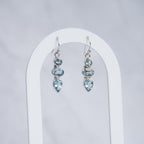 A pair of silver drop earrings with light blue gemstones, displayed on a white arched stand against a light gray background.