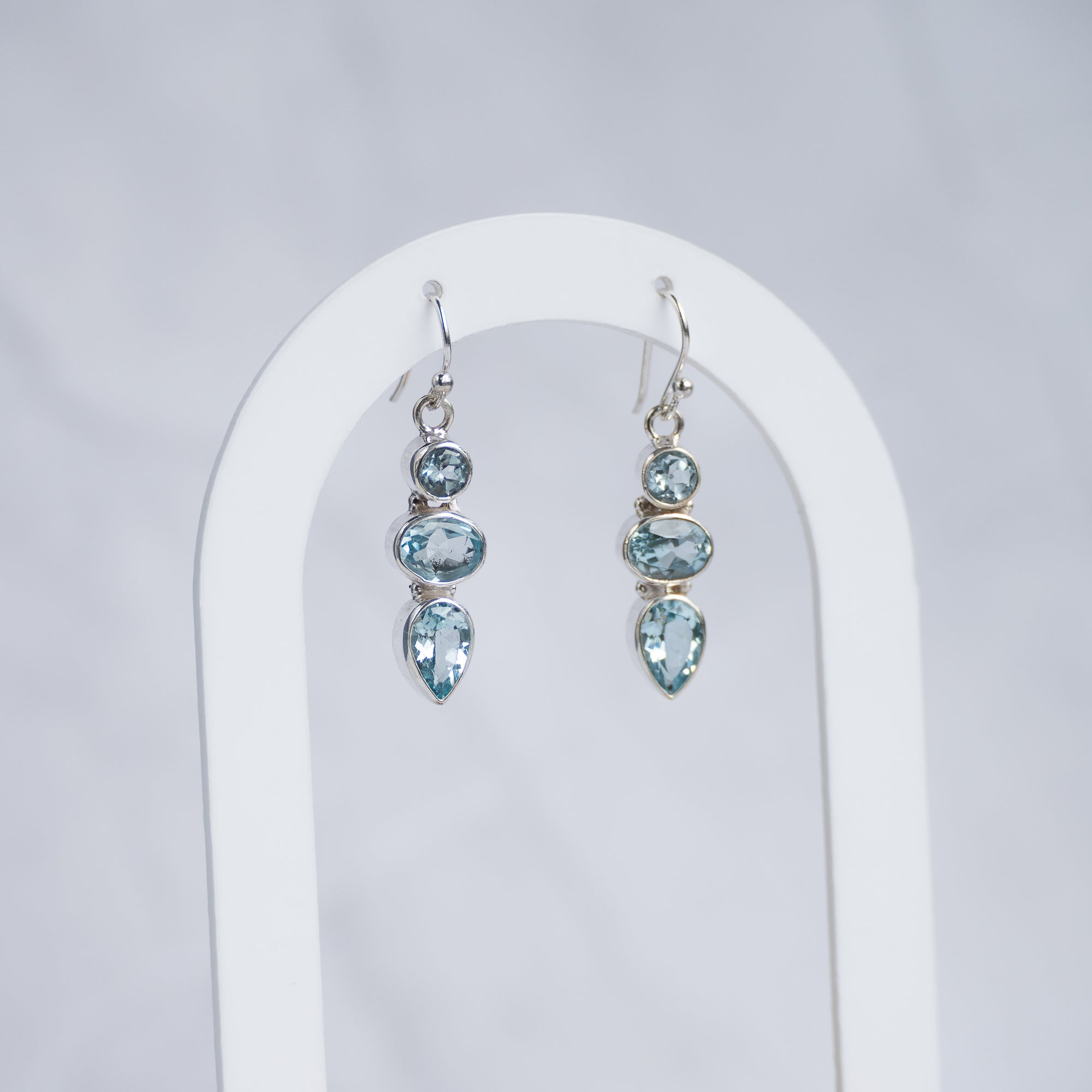 A pair of silver drop earrings with light blue gemstones, displayed on a white arched stand against a light gray background.
