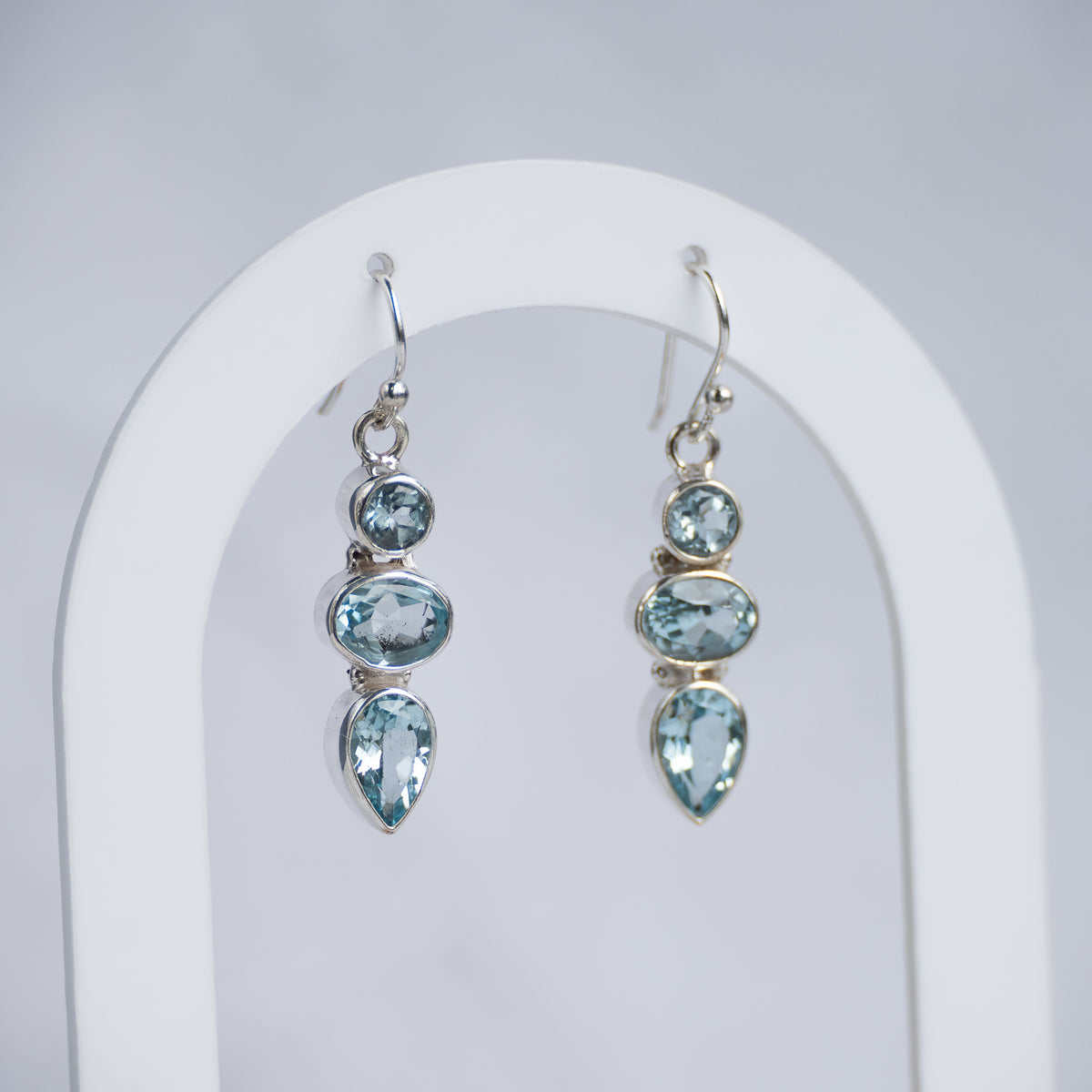 A pair of silver drop earrings with light blue gemstones, displayed on a white arched stand against a light gray background.