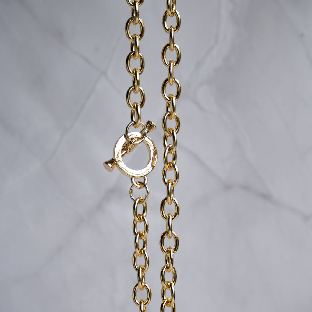 A close-up of a gold chain necklace with a toggle clasp, shown against a light gray marble background.