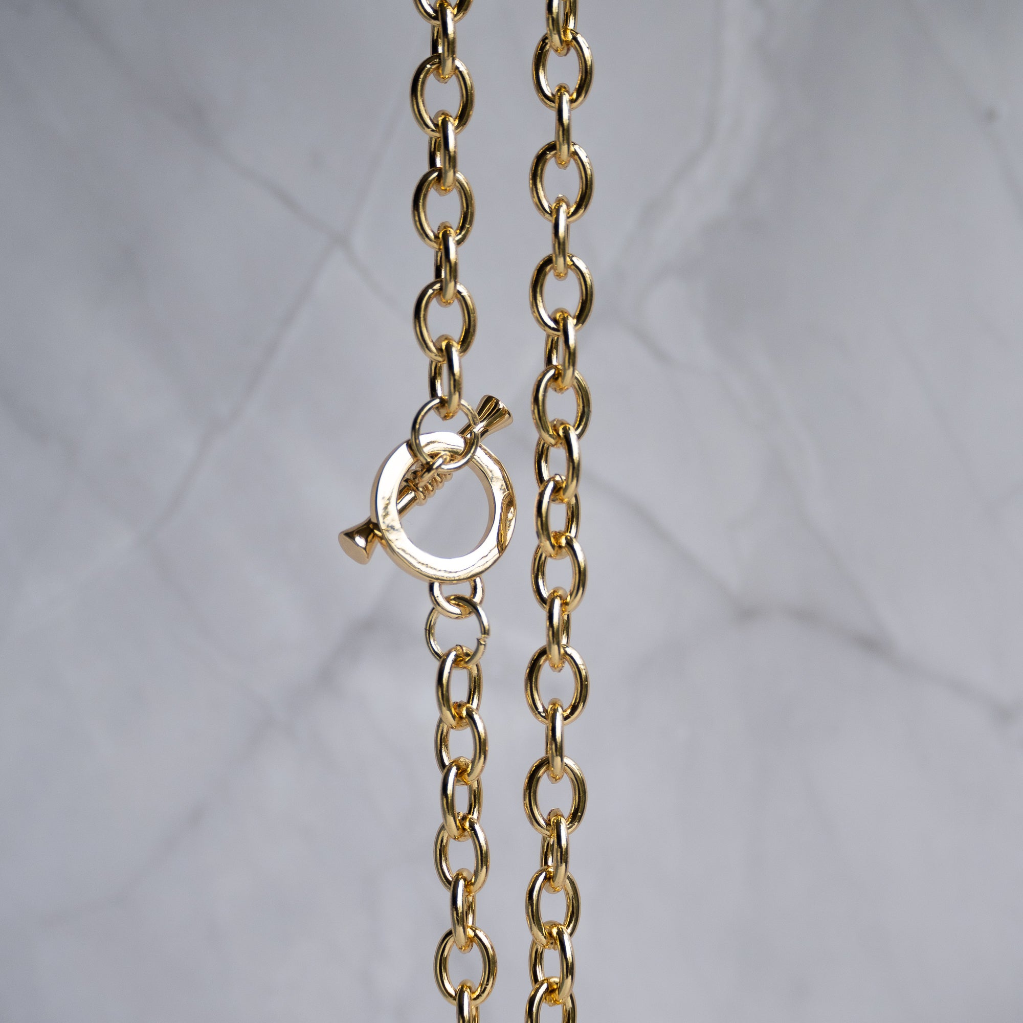 A close-up of a gold chain necklace with a toggle clasp, shown against a light gray marble background.