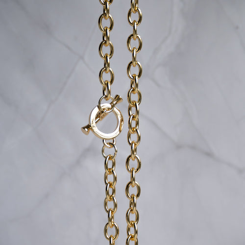 A close-up of a gold chain necklace with a toggle clasp, shown against a light gray marble background.