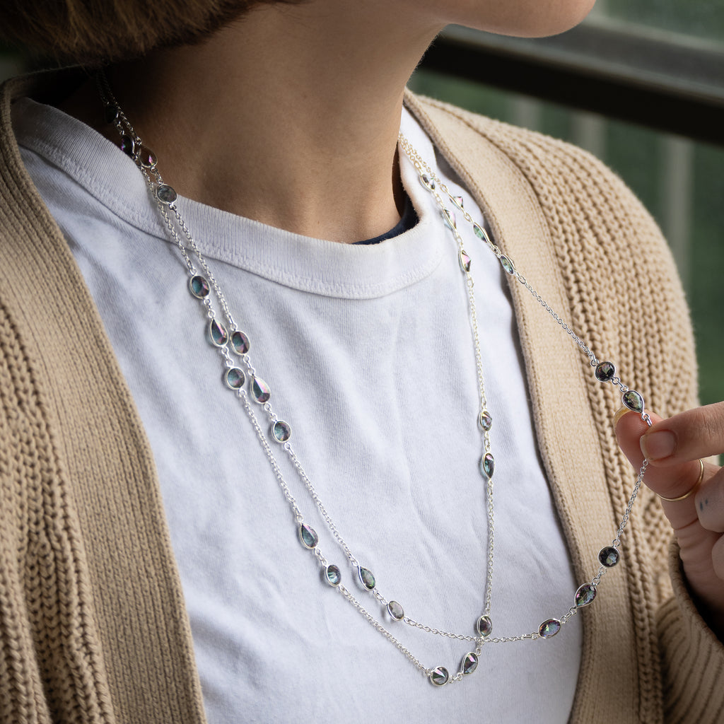 A close-up of a person wearing a layered silver necklace with small dark oval stones over a white t-shirt and beige cardigan.