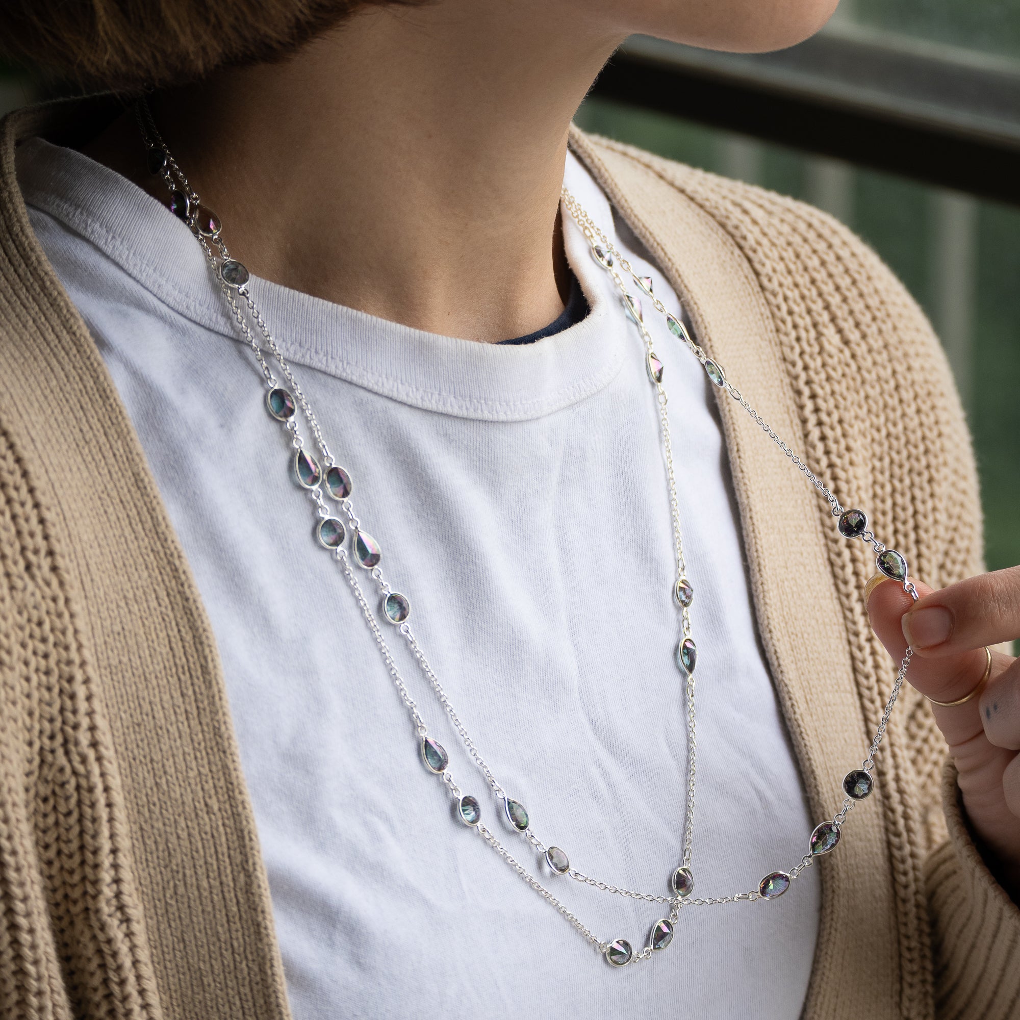 A close-up of a person wearing a layered silver necklace with small dark oval stones over a white t-shirt and beige cardigan.