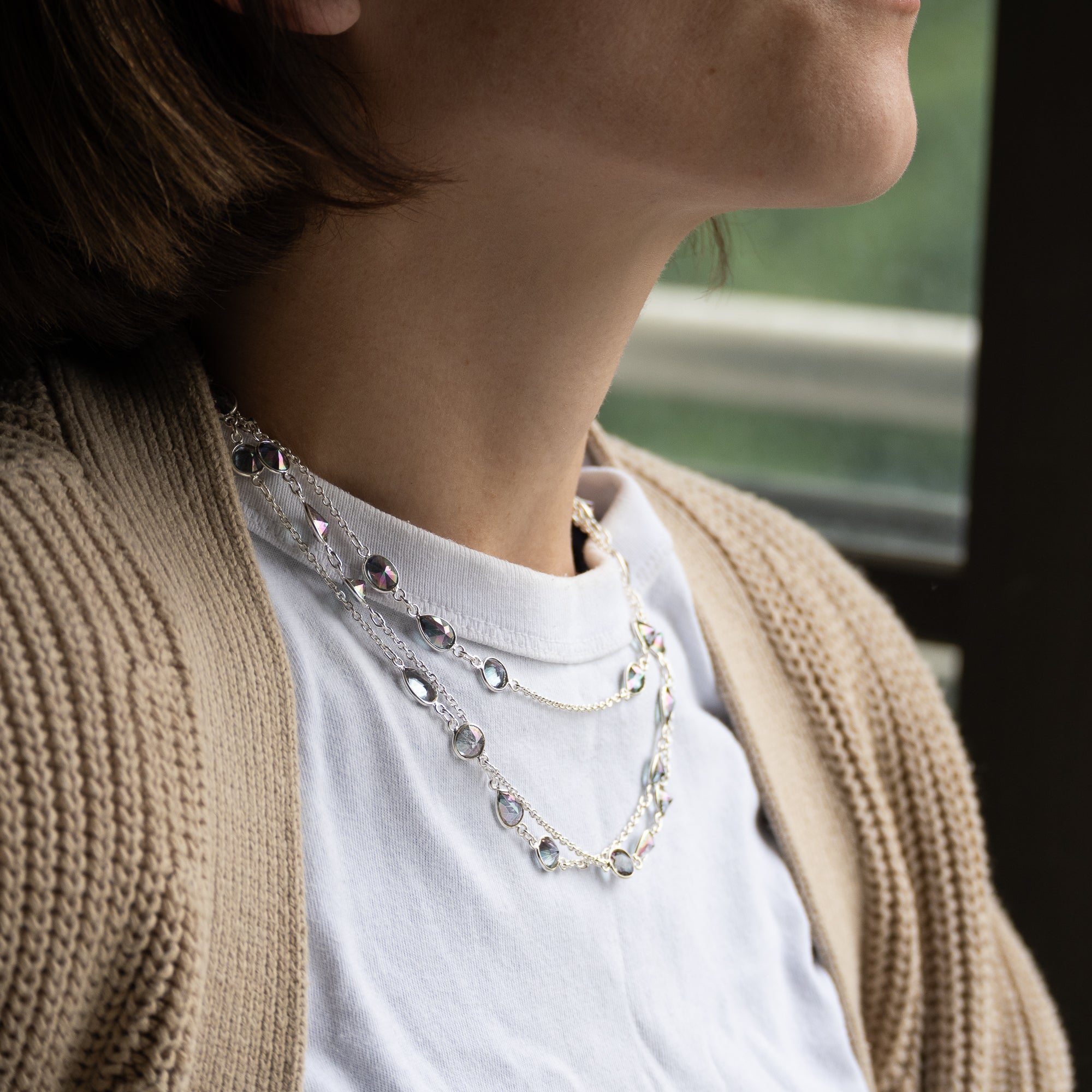 A close-up lifestyle shot of a person wearing a layered silver necklace with clear teardrop crystals and small faceted beads, styled over a white t-shirt and beige cardigan.