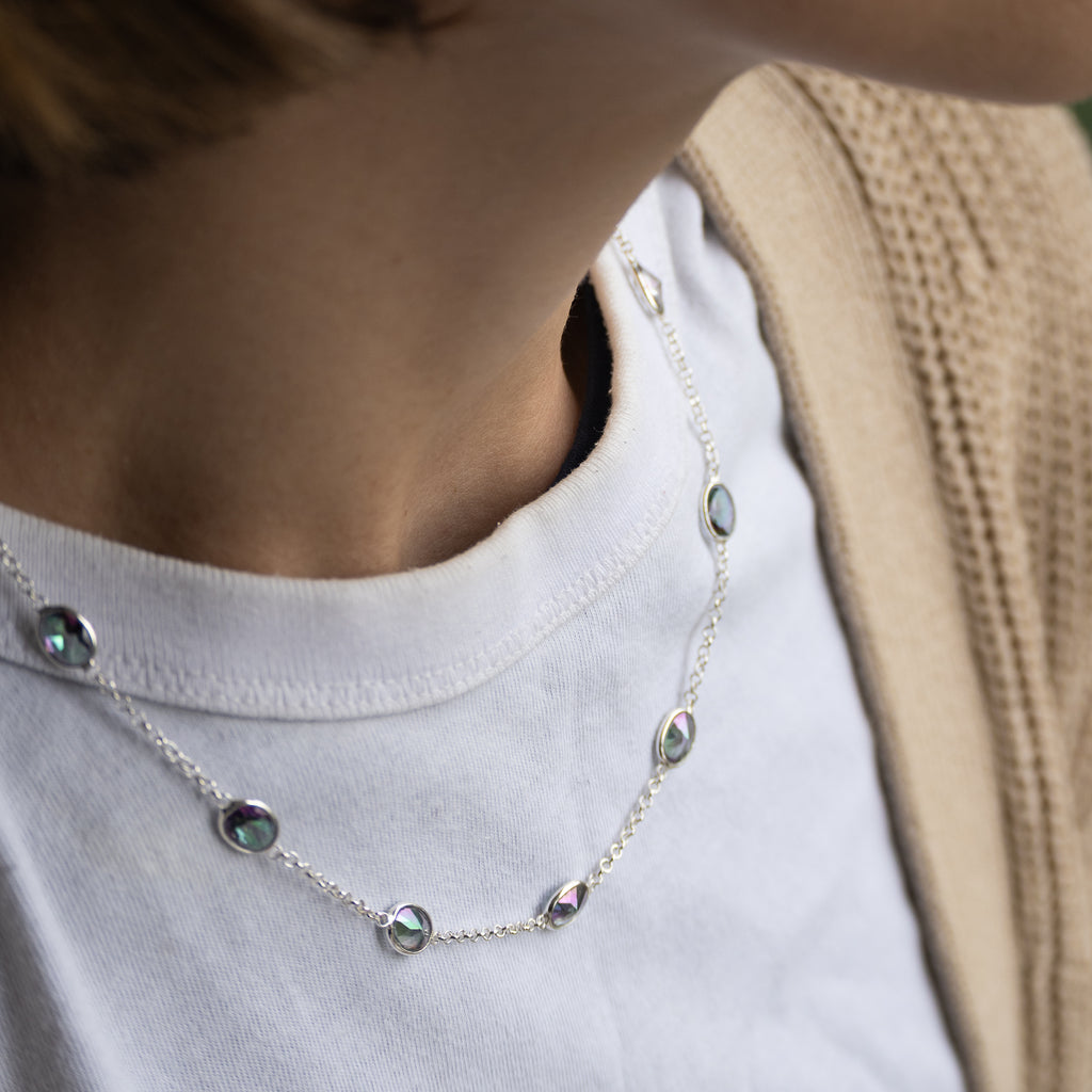 A close-up of a person wearing a delicate silver chain necklace with small oval and teardrop-shaped iridescent stones, styled over a white t-shirt and beige knit cardigan.