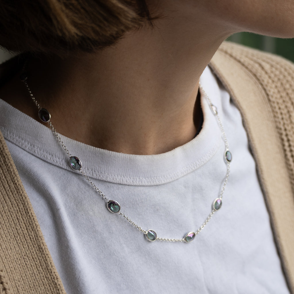 A close-up of a person wearing a delicate silver necklace with small round iridescent beads over a white t-shirt and beige cardigan.