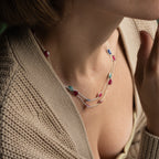 A close-up lifestyle shot of a woman wearing a delicate layered silver necklace adorned with small oval and marquise-cut gemstones in pink, blue, and green, styled with a beige knit cardigan.