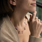 A close-up lifestyle shot of a woman wearing a delicate double-strand necklace with small gold beads, turquoise accents, and red oval gemstones, styled with a beige knit cardigan.