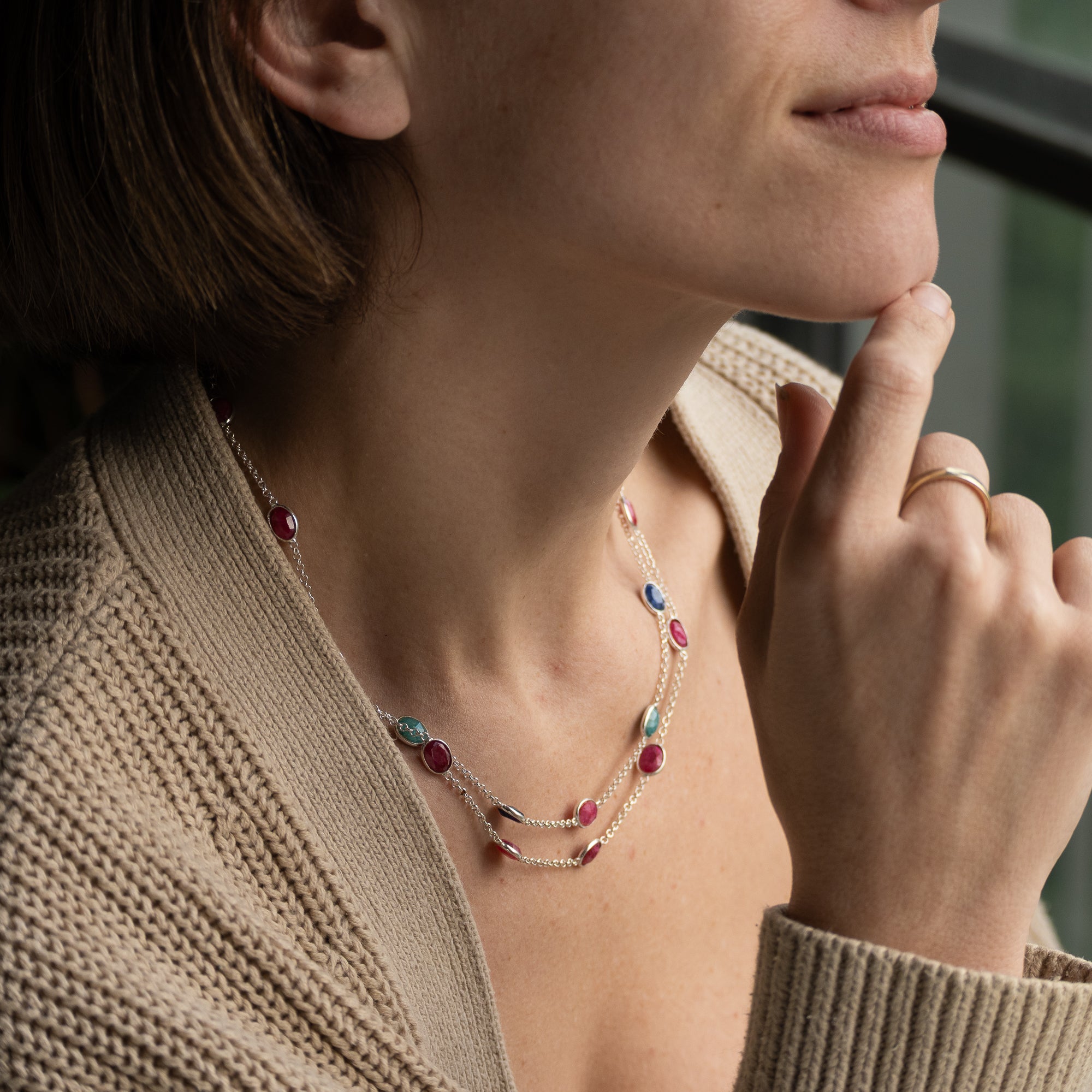 A close-up lifestyle shot of a woman wearing a delicate double-strand necklace with small gold beads, turquoise accents, and red oval gemstones, styled with a beige knit cardigan.