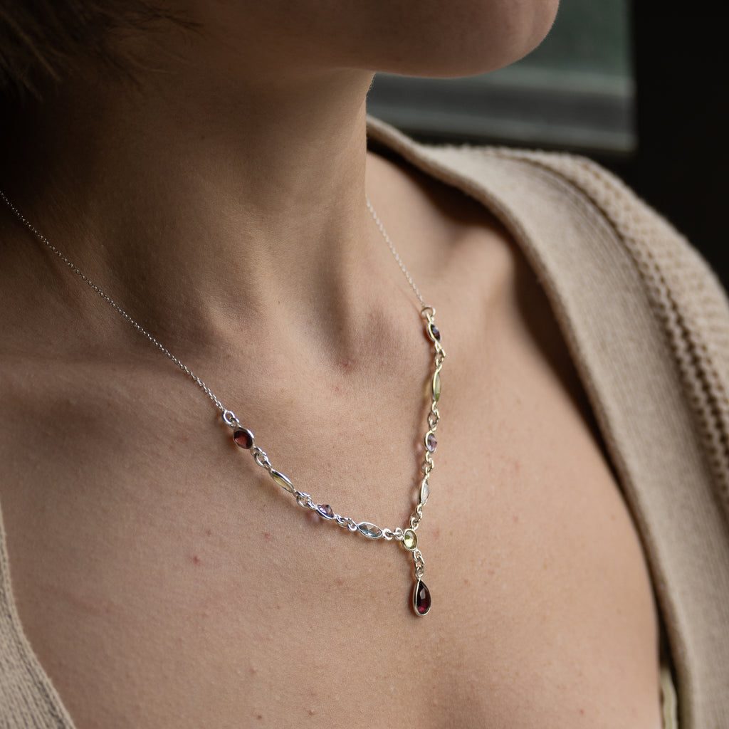 A delicate silver-toned necklace with a Y-shaped design, featuring small clear faceted stones and two dark red teardrop gemstones, shown worn on a model's neck and chest.
