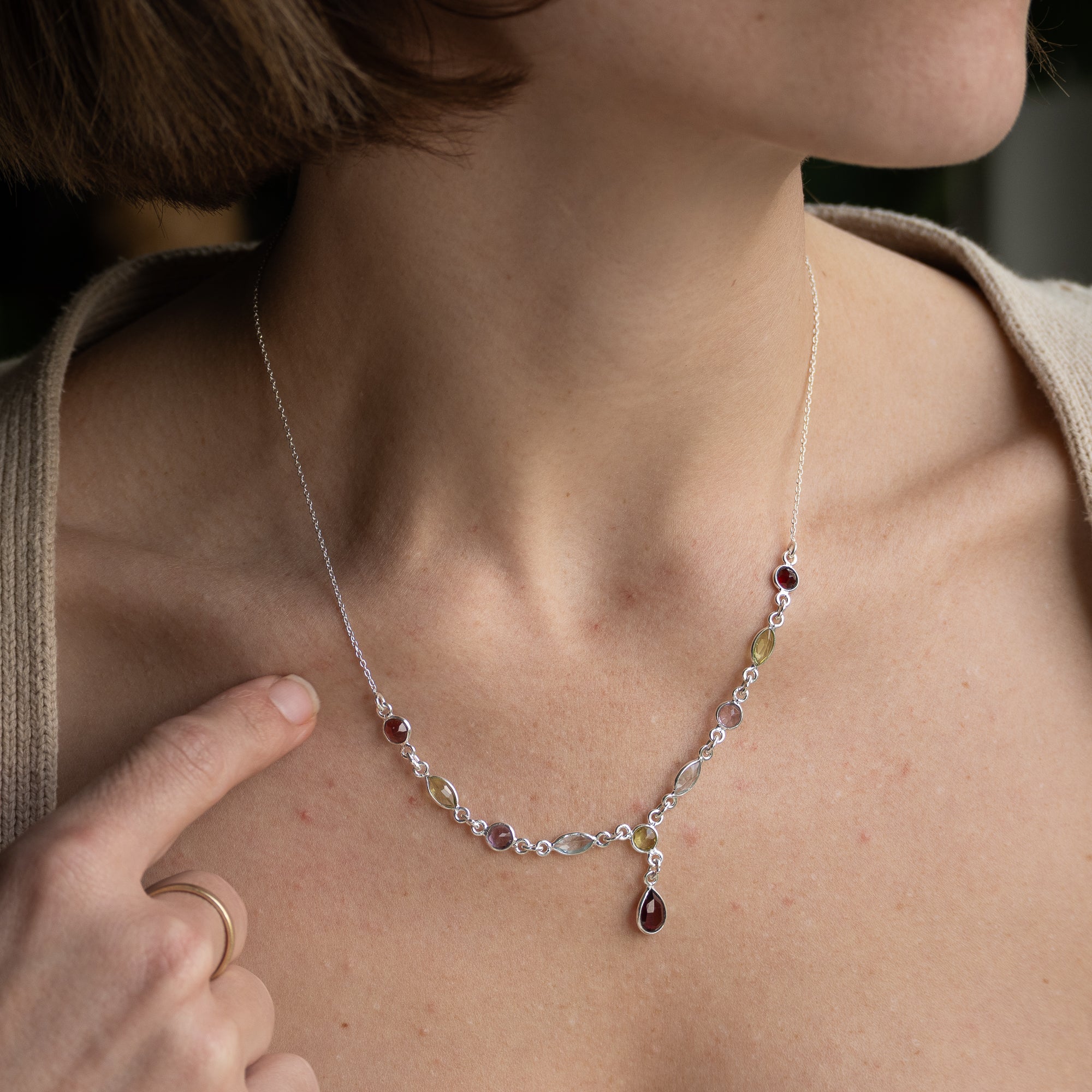A close-up of a woman wearing a delicate silver-toned necklace with small clear stones, burgundy gemstone accents, and a teardrop-shaped dark red pendant.