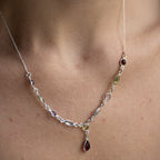 A close-up of a delicate silver necklace worn on a person's neck, featuring small oval and marquise-cut gemstones in silver settings with a teardrop-shaped dark red gemstone pendant.