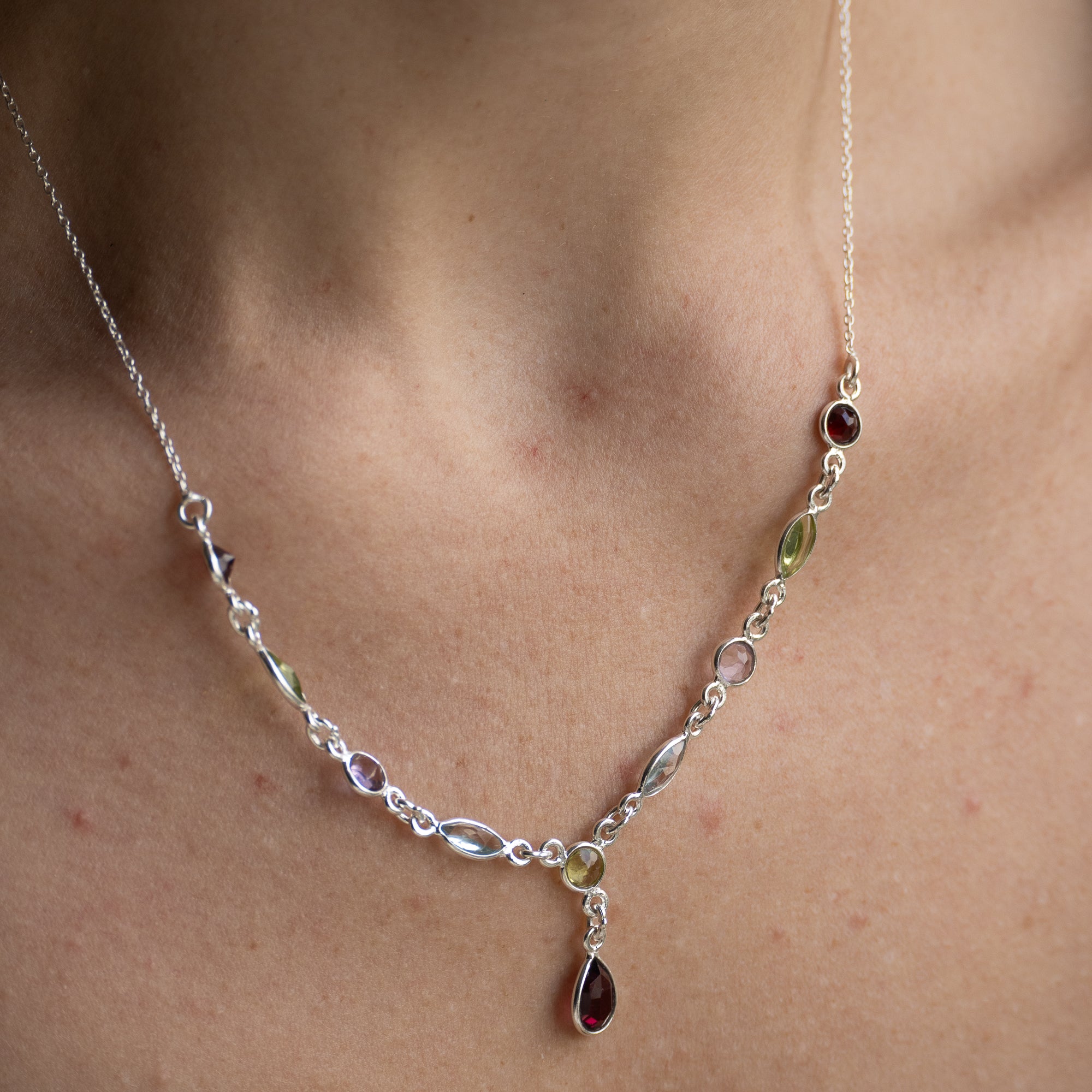 A close-up of a delicate silver necklace worn on a person's neck, featuring small oval and marquise-cut gemstones in silver settings with a teardrop-shaped dark red gemstone pendant.