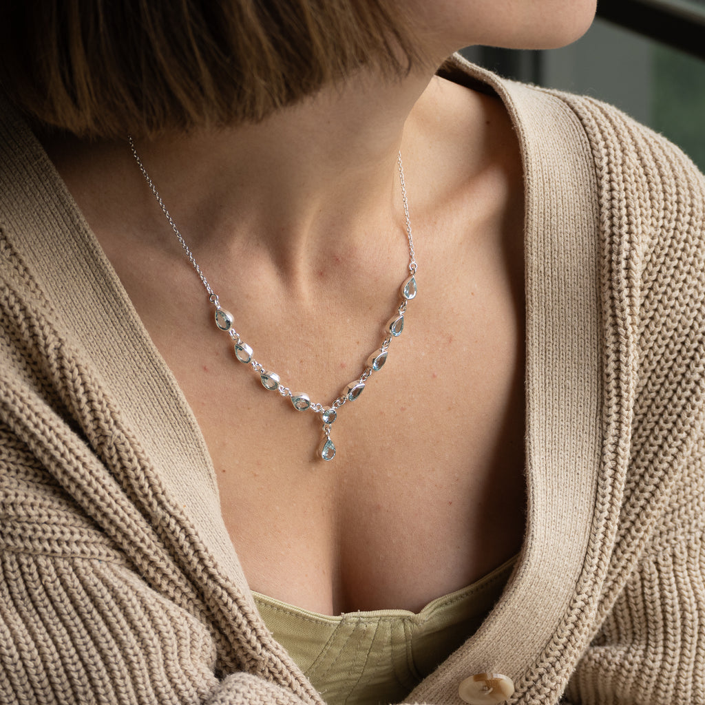 A close-up of a woman wearing a delicate silver necklace with clear teardrop-shaped crystals and a central teardrop pendant, styled with a beige cardigan.