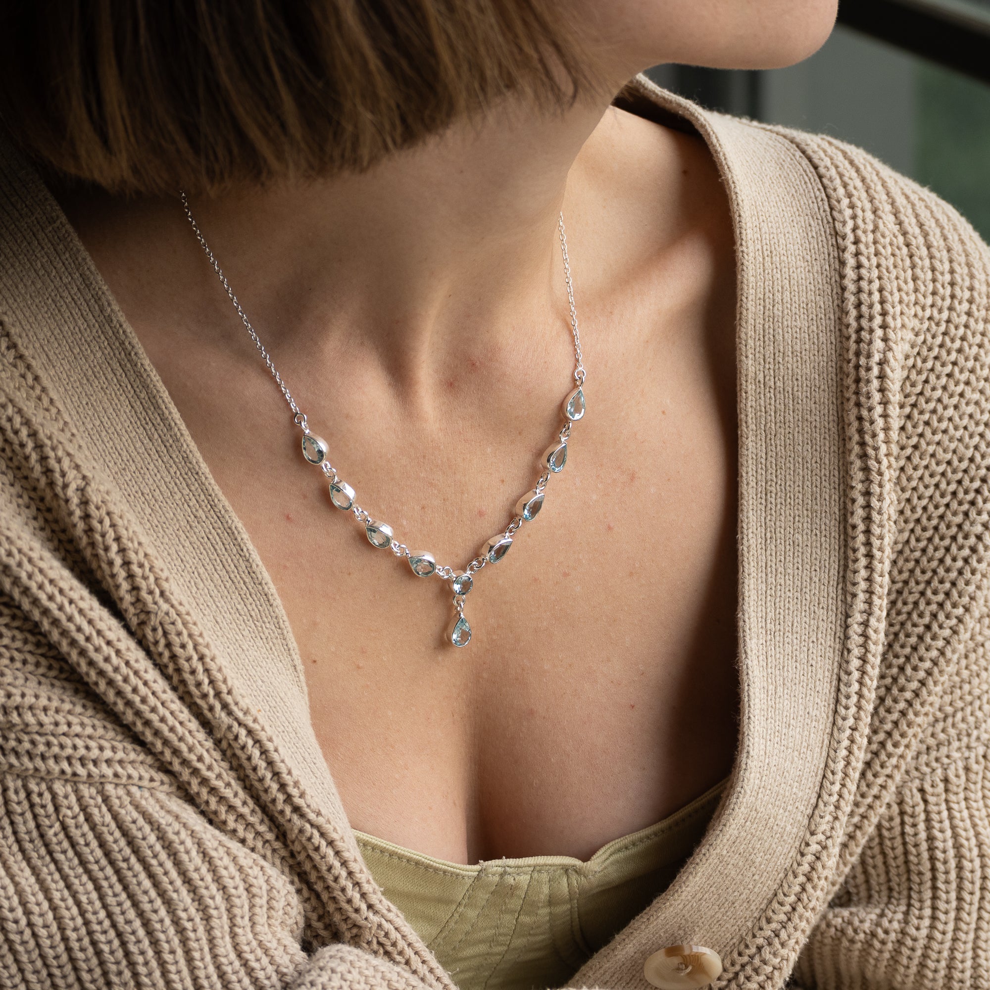 A close-up of a woman wearing a delicate silver necklace with clear teardrop-shaped crystals and a central teardrop pendant, styled with a beige cardigan.