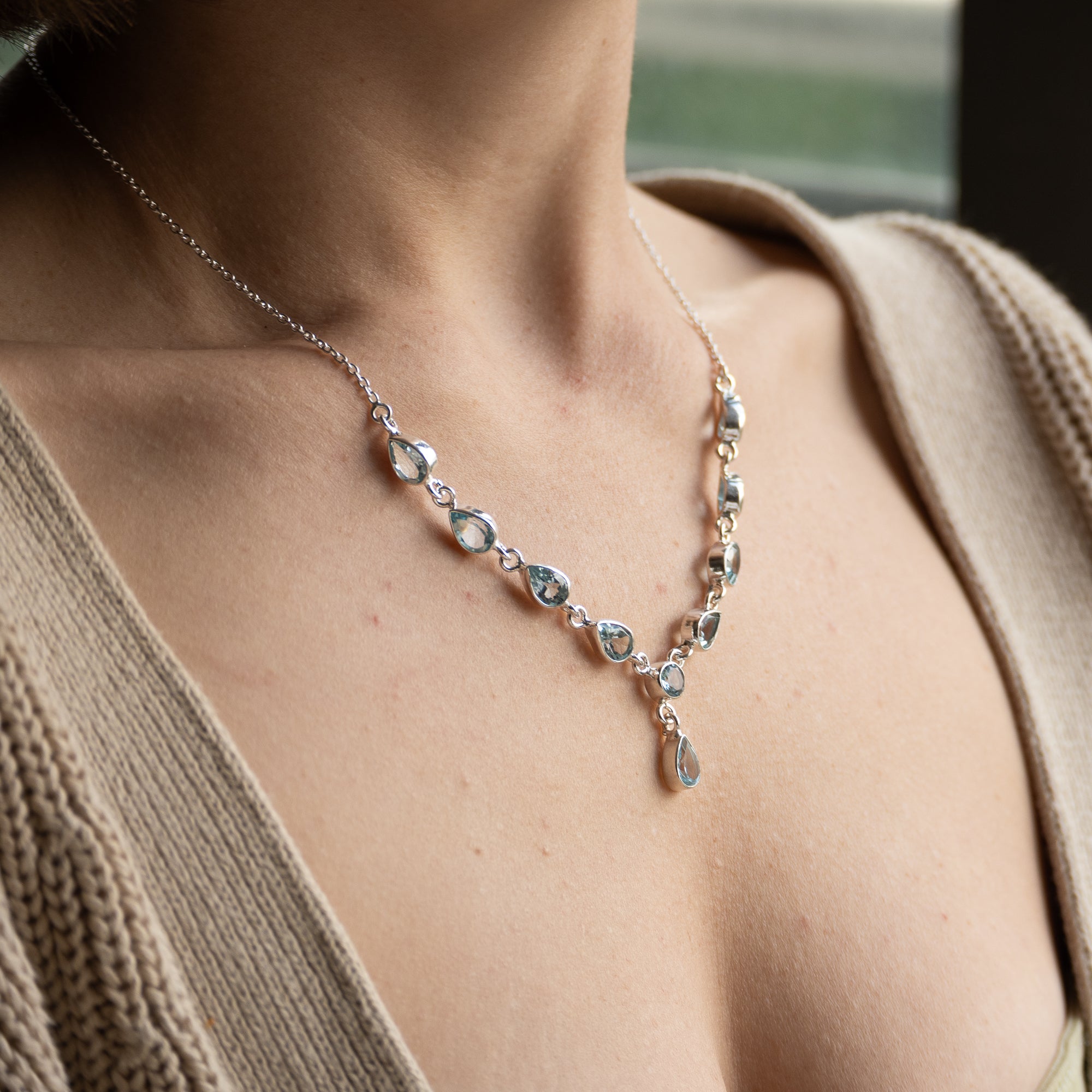 A close-up of a model wearing a delicate silver necklace with clear faceted gemstones and a small teardrop pendant, styled with a beige knit cardigan.