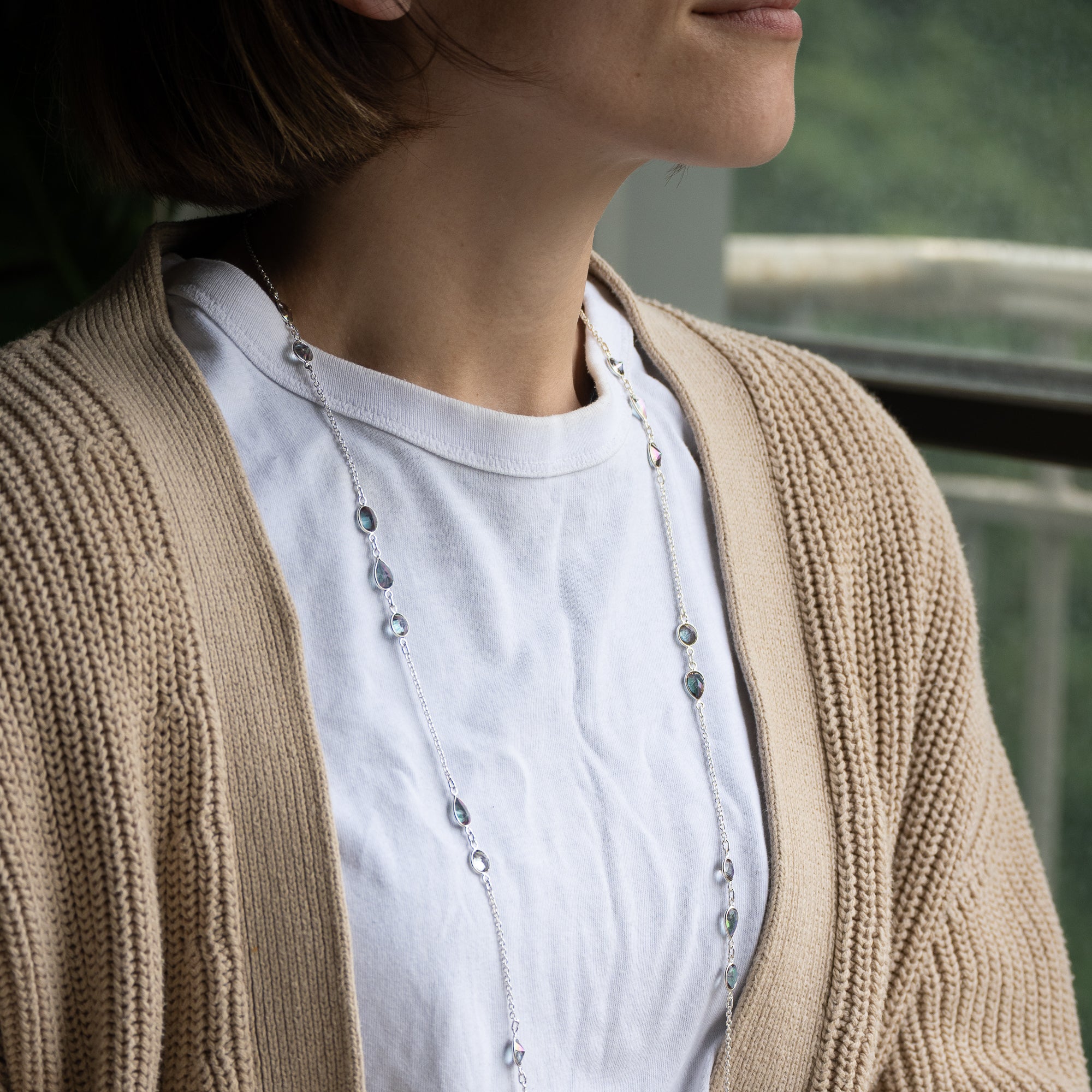 A close-up lifestyle shot of a person wearing a delicate silver beaded necklace over a white t-shirt and a beige cable-knit cardigan.