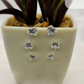 A pair of sterling silver creeper earrings with cubic zirconia flowers displayed on a white surface, partially in front of a green potted plant.