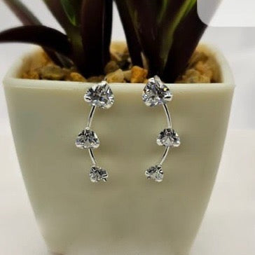 A pair of sterling silver creeper earrings with cubic zirconia flowers displayed on a white surface, partially in front of a green potted plant.