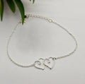 A sterling silver bracelet featuring two interlinked heart-shaped charms, displayed against a white background with a plant in the background.