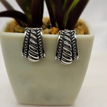 A pair of sterling silver patterned drop earrings displayed in a white vase with a green plant background.
