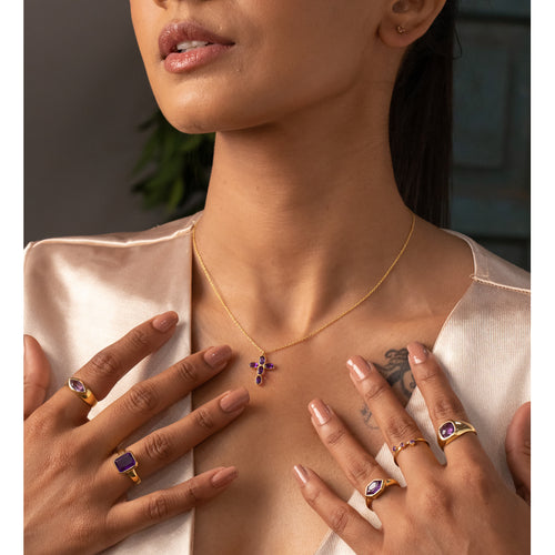 Woman wearing a gold necklace with a purple pendant, surrounded by multiple rings on her fingers.
