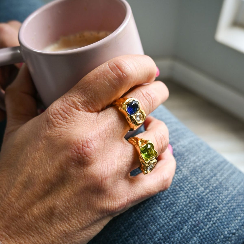 Hand holding a pink mug with a ring featuring gemstones, sitting on a blue cushion.