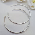 A pair of 50 mm sterling silver hoop earrings displayed against a white background with two white flowers.