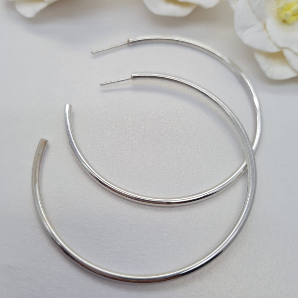 A pair of 50 mm sterling silver hoop earrings displayed against a white background with two white flowers.