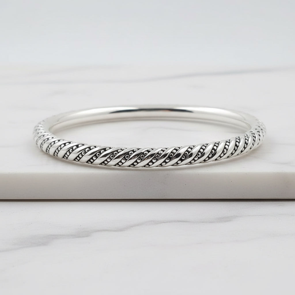 A sterling silver hollow bracelet with a geometric spiral pattern, displayed on a white surface next to a green plant.