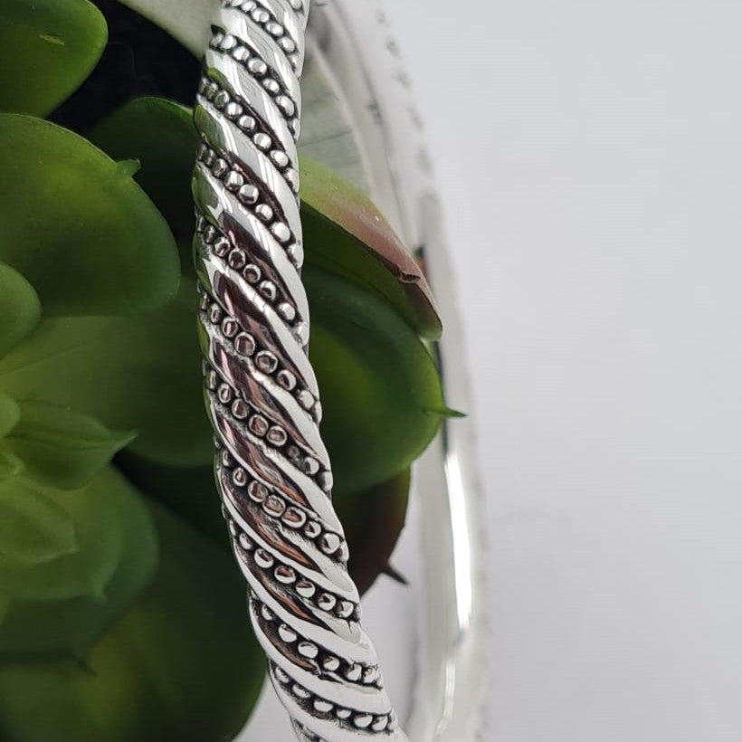 A sterling silver hollow bracelet with a geometric spiral pattern, displayed on a white surface next to a green plant.
