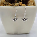 A pair of sterling silver bee earrings dangle over a white ceramic pot with plants in the background.