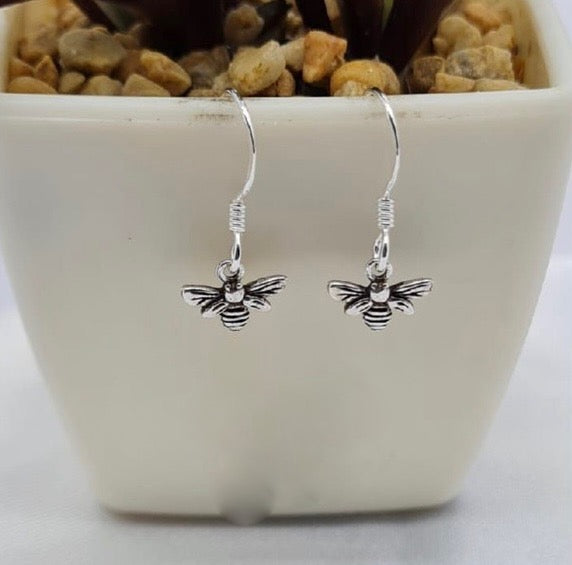 A pair of sterling silver bee earrings dangle over a white ceramic pot with plants in the background.