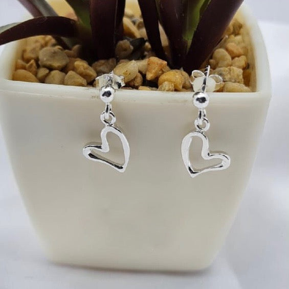 A pair of sterling silver dangling heart-shaped earrings displayed in a white vase with a plant background.