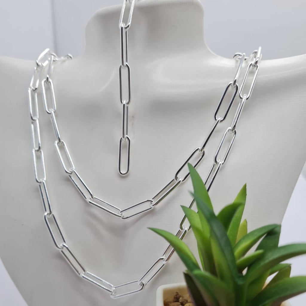 A sterling silver paperclip chain necklace displayed on a white stand, with a few paperclips varying in length, next to a small potted plant.