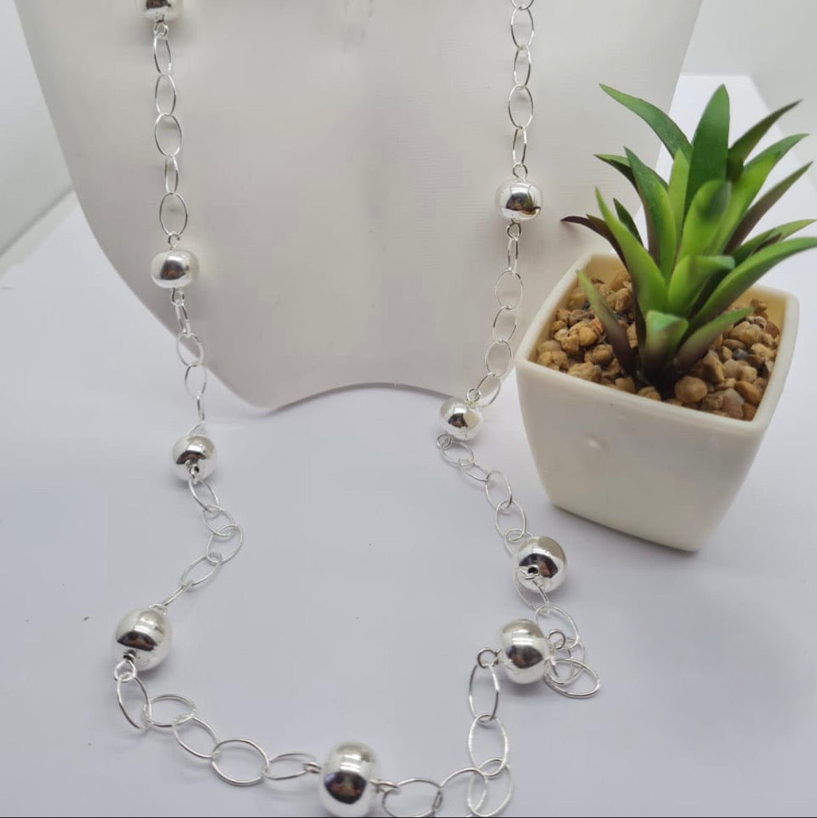 A 70 cm long silver ball chain necklace displayed on a mannequin bust, with a small potted plant beside it.